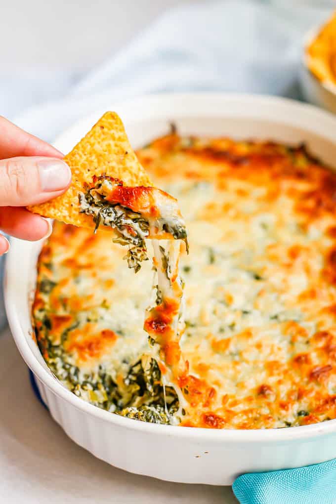 A hand scooping healthy spinach dip from a serving dish with a tortilla chip