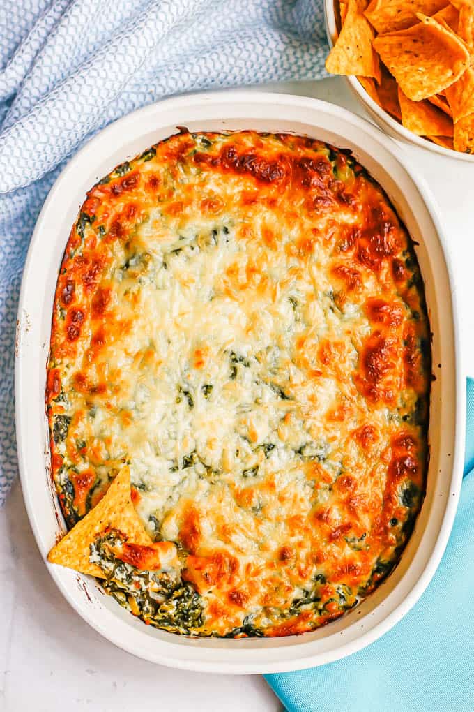 A baked spinach dip with a cheesy browned crust and a bowl of chips nearby for dipping