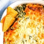 A browned and cheesy baked spinach dip in a white casserole dish with tortilla chips tucked into the corner for dipping and a text overlay on the photo.