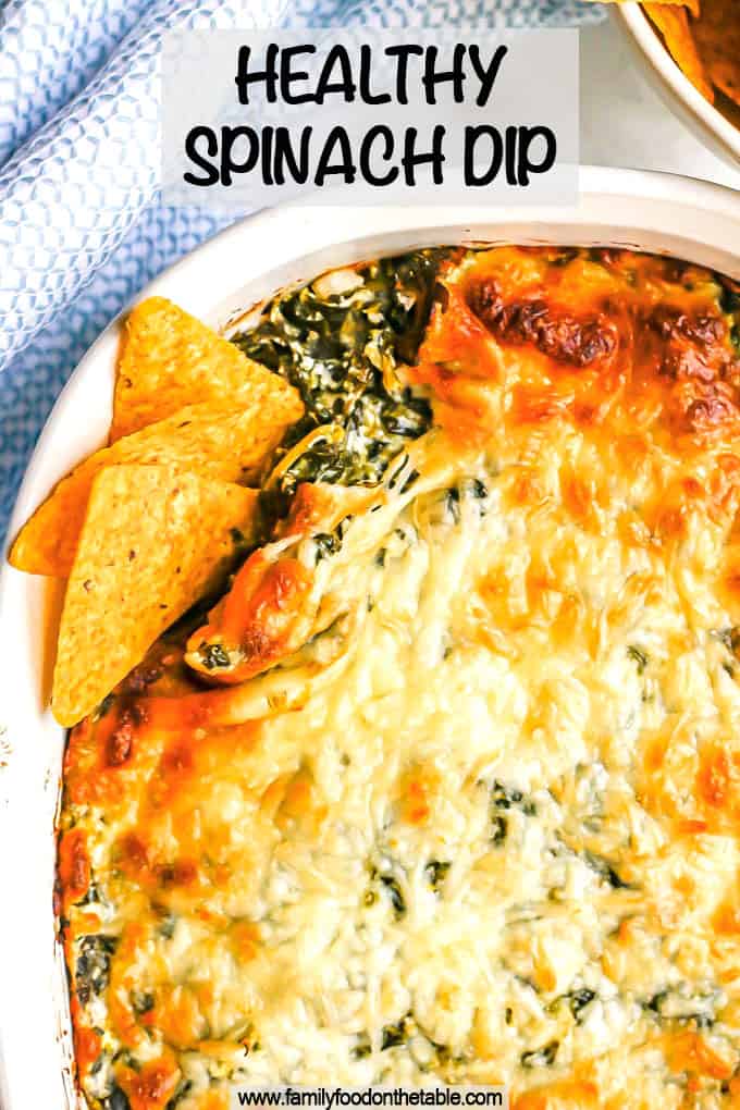 A browned and cheesy baked spinach dip in a white casserole dish with tortilla chips tucked into the corner for dipping and a text overlay on the photo