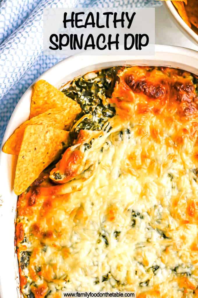 A browned and cheesy baked spinach dip in a white casserole dish with tortilla chips tucked into the corner for dipping and a text overlay on the photo.