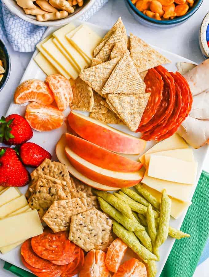 A white platter filled with cheese, crackers, cured meats, fruits and snacks for kids