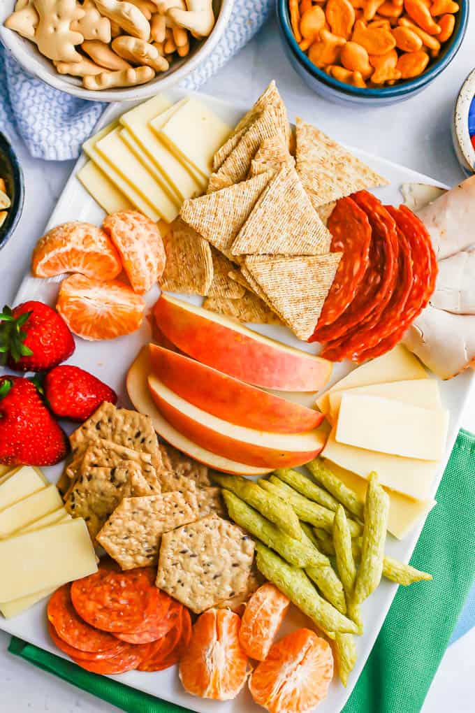 A white platter filled with cheese, crackers, cured meats, fruits and snacks for kids