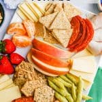 A white platter filled with cheese, crackers, cured meats, fruits and snacks for kids with a text overlay on the photo