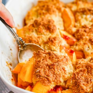 A serving spoon lifting a scoop of fresh peach cobbler out of a white casserole dish