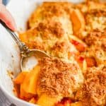 A serving spoon lifting a scoop of fresh peach cobbler out of a white casserole dish and a text overlay on the photo