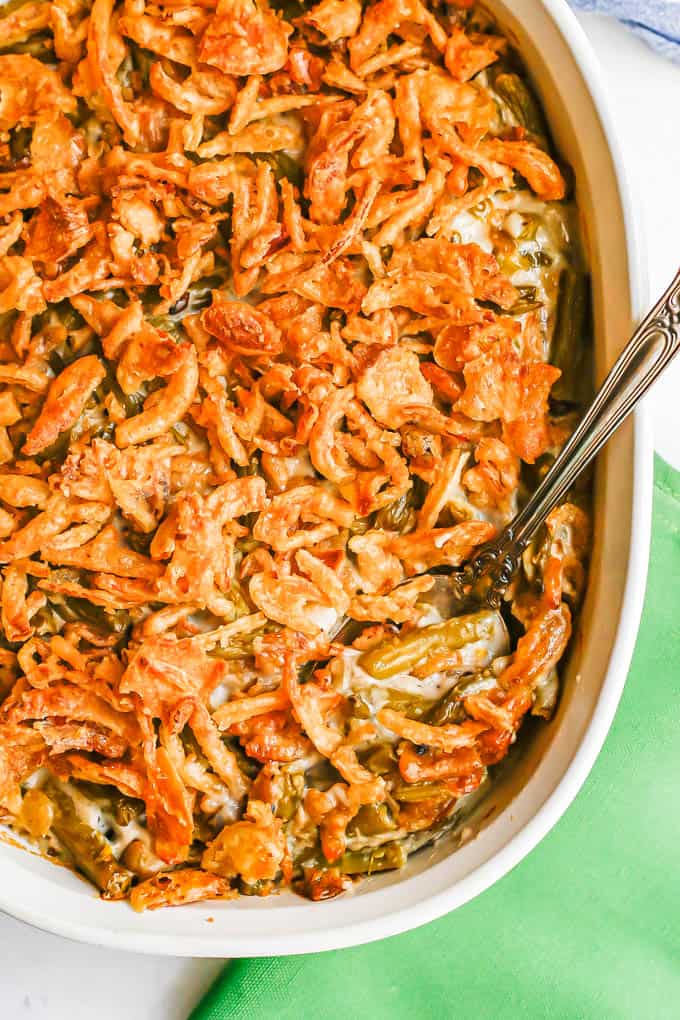 Green bean casserole with French fried onions on top in a white oval baking dish after cooking and a serving spoon tucked in the side