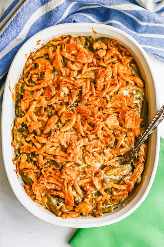 Green bean casserole with French fried onions on top in a white oval baking dish after cooking with a serving spoon in the side and green napkins nearby