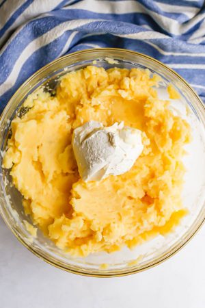 Mashed cooked potatoes in a glass bowl with cream cheese dolloped on top before being mixed in