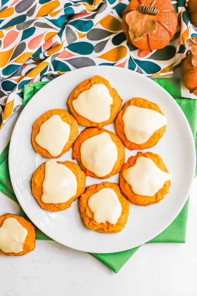 Overhead shot of a white plate with pumpkin cookies on top of green napkins with a colorful towel and some small decorative pumpkins to the back