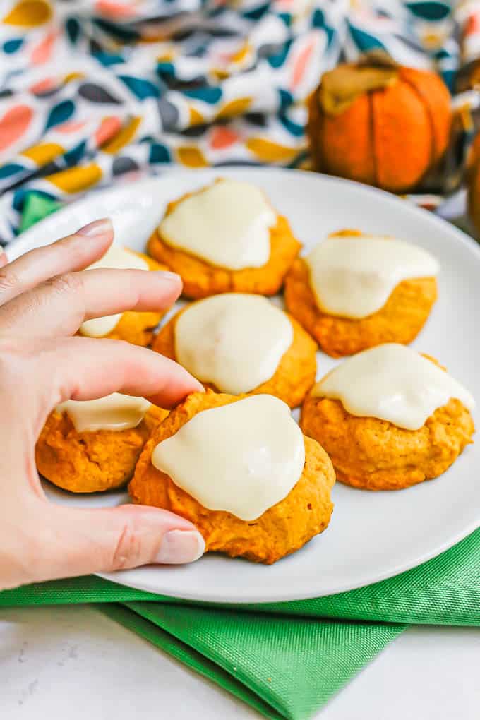 A hand about to pick up a cookie from a white plate full of soft pumpkin cookies with orange icing on top