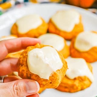 A hand holding a small pumpkin cookie with orange icing up from a white plate