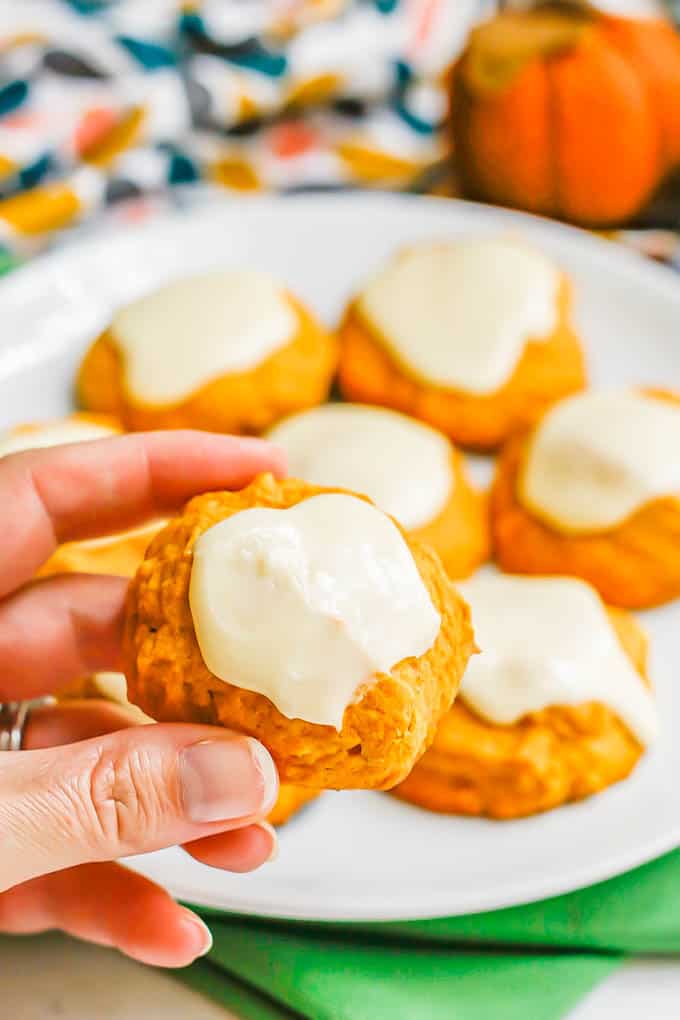 A hand holding a small pumpkin cookie with orange icing up from a white plate