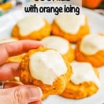 A hand holding a small pumpkin cookie with orange icing up from a white plate with a text overlay on the photo
