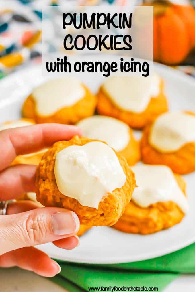 A hand holding a small pumpkin cookie with orange icing up from a white plate with a text overlay on the photo