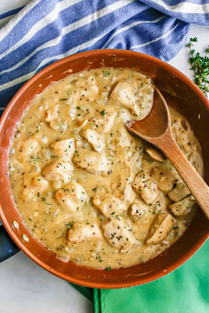 A copper skillet with cubed chicken in gravy with a wooden spoon resting in the skillet and a blue striped towel and green napkins nearby