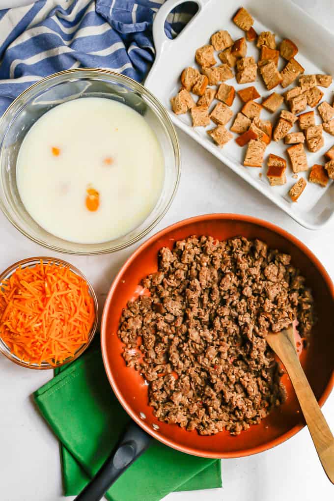 Ingredients laid out for making a breakfast casserole, including a pan of cooked, crumbled sausage, a bowl of shredded cheese, a bowl with eggs and milk and a casserole dish with cubed bread.