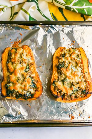 Two halves of a butternut squash stuffed with sausage and spinach and topped with Parm cheese on a foil lined baking sheet