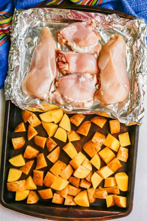 Chicken breasts and thighs in a foil pack beside potatoes on a sheet pan before being cooked