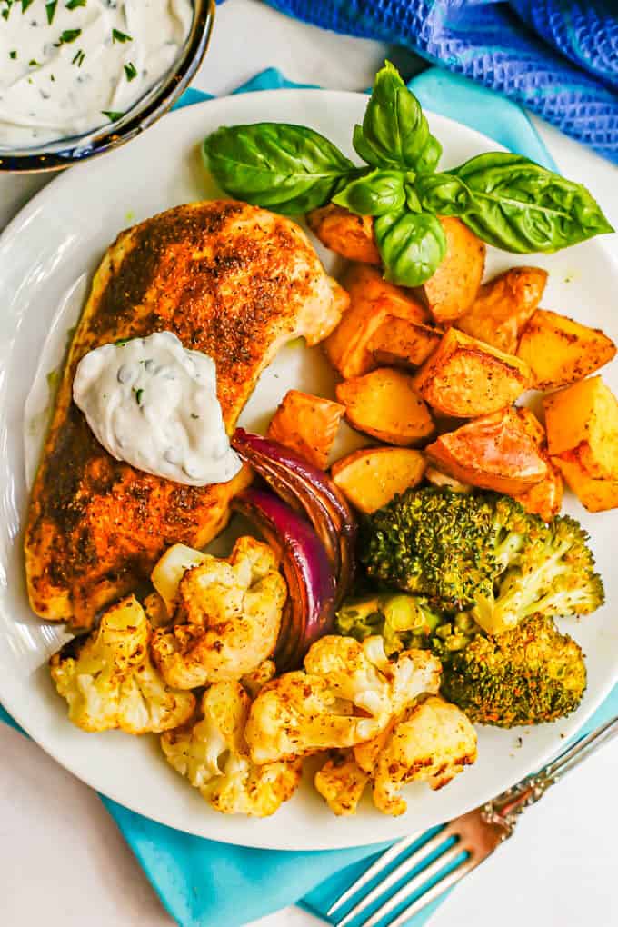 A white dinner plate with roasted chicken and veggies served with a basil sprig and an herbed yogurt sauce