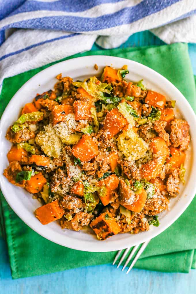 A white bowl with turkey sausage, sweet potatoes and Brussels sprouts topped with Parmesan cheese
