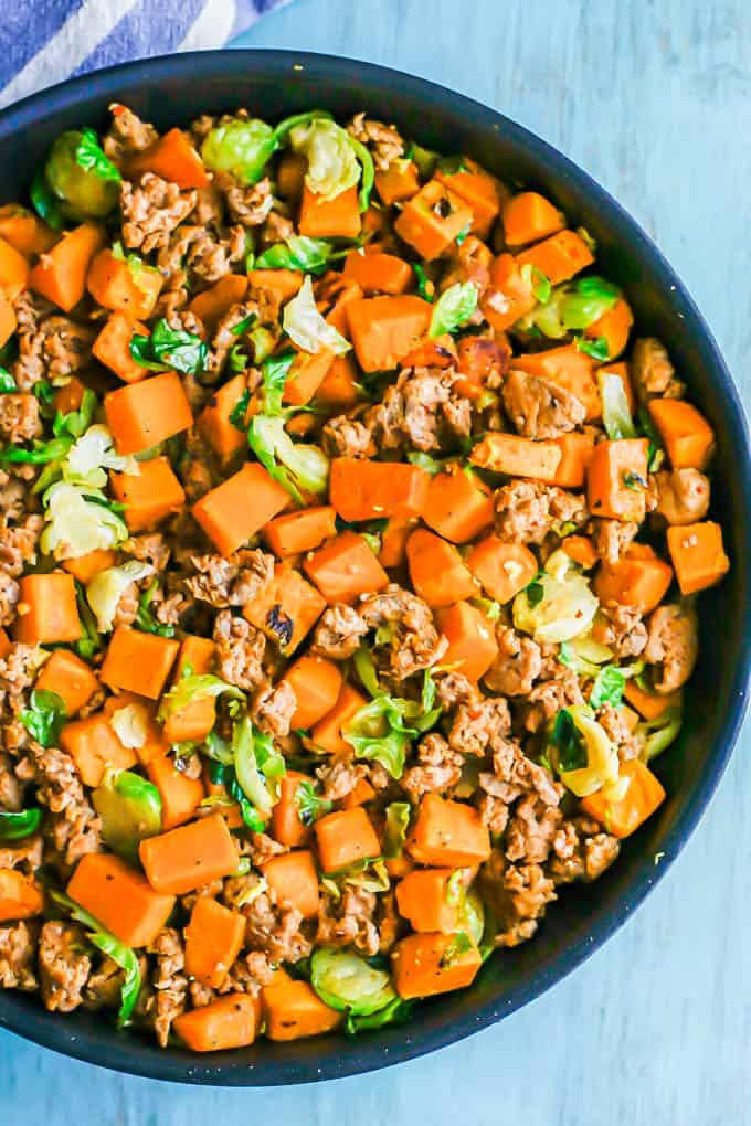 A large dark skillet with a cooked mixture of sausage, sweet potatoes and Brussels sprouts