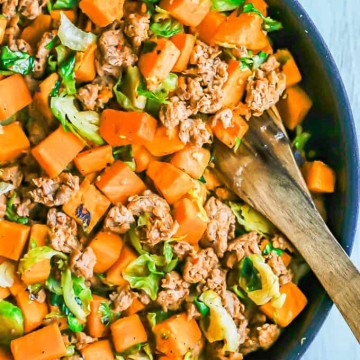 A large skillet with turkey sausage, sweet potatoes and Brussels sprouts with a wooden spoon resting in the pan
