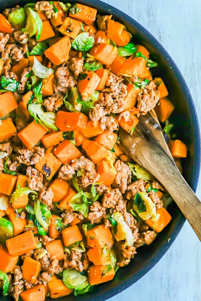 A large skillet with turkey sausage, sweet potatoes and Brussels sprouts with a wooden spoon resting in the pan