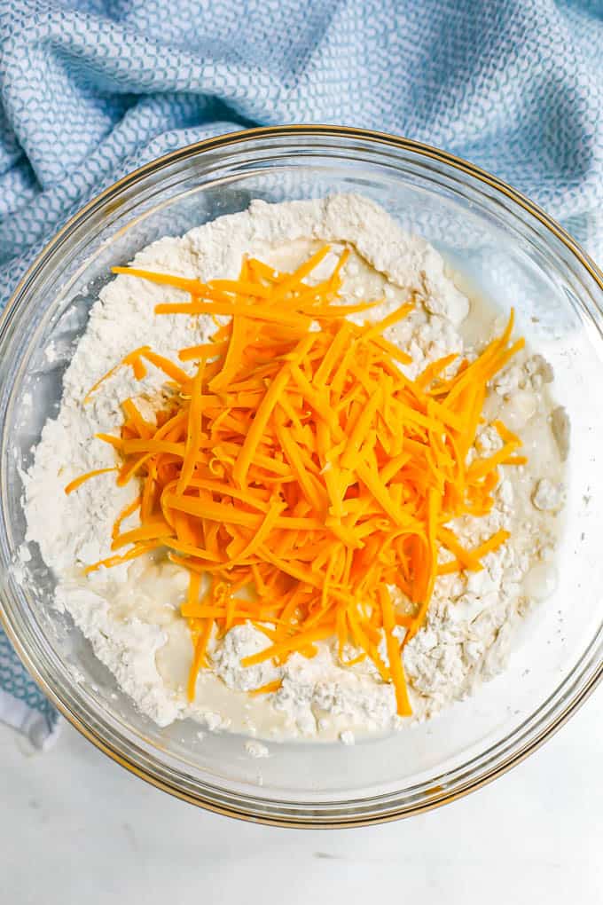 A glass bowl with a dry flour mixture topped with milk and shredded cheddar cheese before it's mixed in