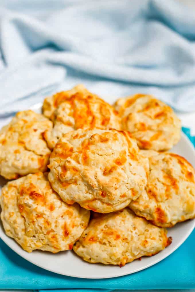 A white serving plate of cheddar garlic drop biscuits set on a couple of turquoise napkins with a kitchen towel in the background