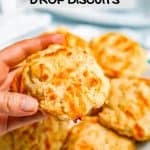 A hand holding up a cheddar garlic drop biscuit from a plate with a text overlay on the photo