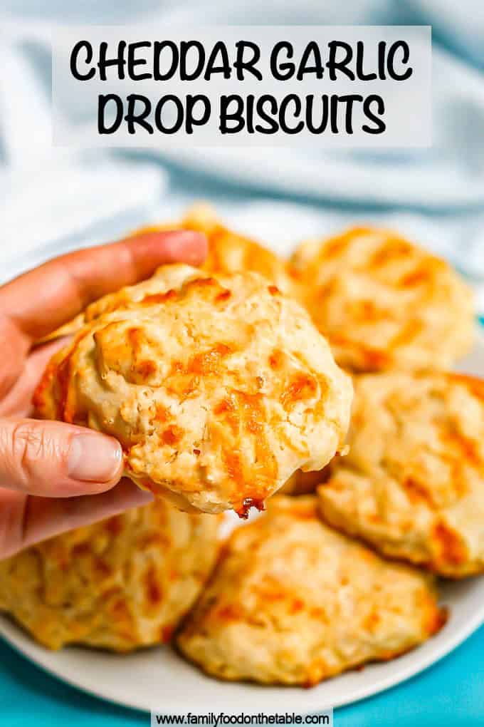 A hand holding up a cheddar garlic drop biscuit from a plate with a text overlay on the photo