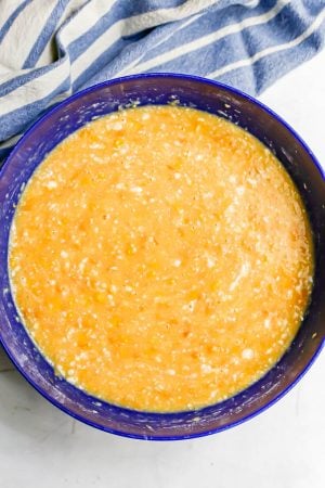 A large blue bowl with a liquidy batter for corn pudding casserole and a blue striped towel in the background