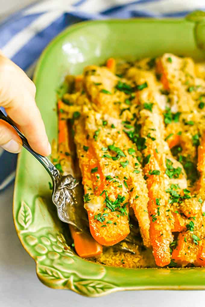 A silver serving fork lifting up some roasted and coated carrots topped with parsley from a green baking dish