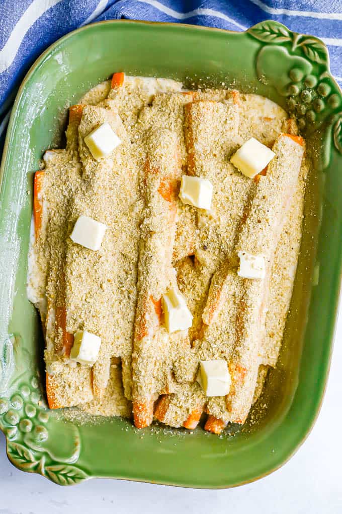 Long carrot strips in a green baking dish, topped with breadcrumbs and dots of butter before being baked