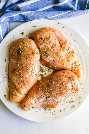 Three raw chicken breasts on a large white plate with salt, pepper and finely chopped fresh rosemary sprinkled on for seasoning