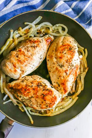 Three seasoned and seared chicken breasts in a large skillet with sliced onions