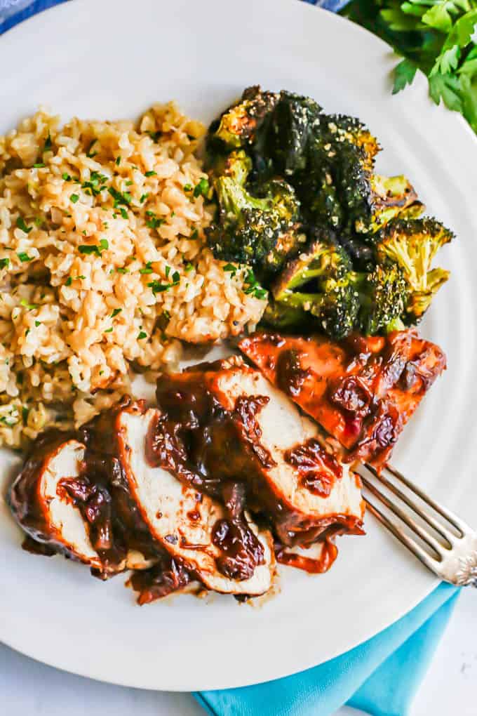 Overhead shot of sliced cranberry balsamic chicken breasts on a white dinner plate with rice pilaf and roasted broccoli