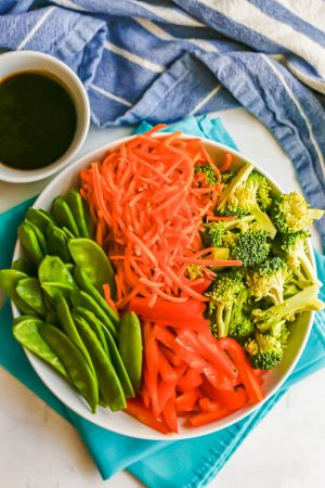 A white bowl with fresh veggies including broccoli, carrots, snow peas and red onion with a white bowl of sauce to the side