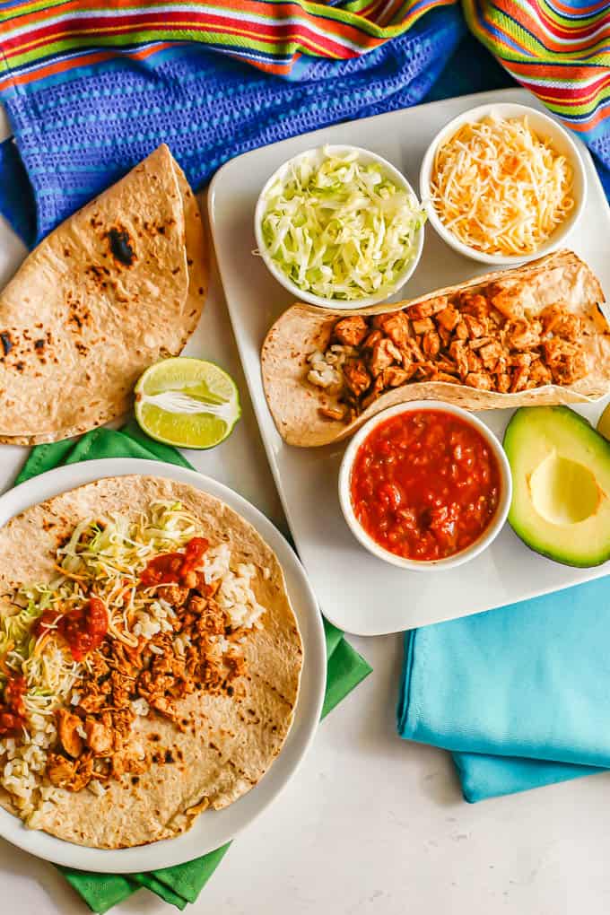 A plate with an assembled taco and a plate with a folded taco and bowls of taco toppings plus colorful towels and napkins with the plates