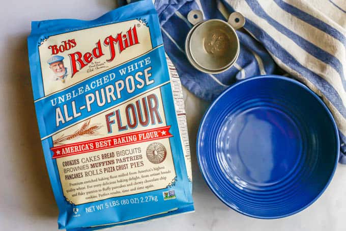 A bag of Bob's Red Mill all-purpose flour beside a blue bowl and some measuring cups with a blue striped towel nearby
