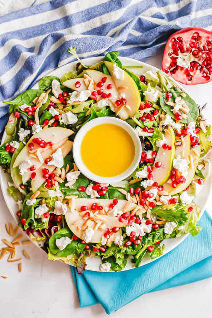 A large round white platter with a Christmas salad of mixed greens, pears, pomegranates and a citrus dressing in a small bowl in the middle