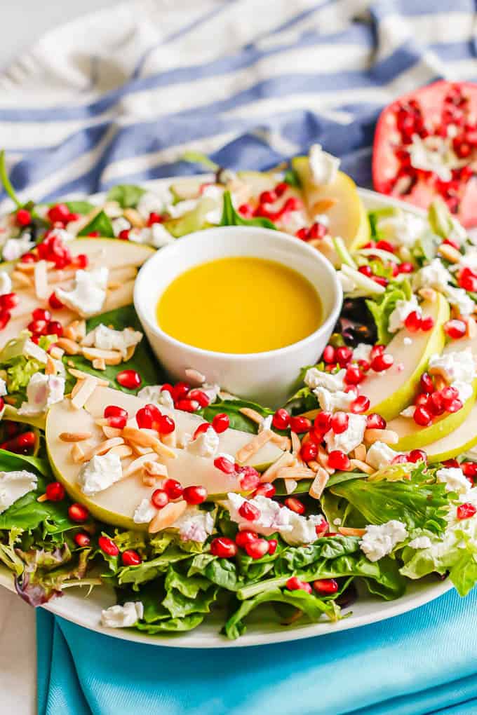 A mixed greens salad with pears, pomegranates, slivered almonds and goat cheese served on a large white platter with citrus dressing in the middle