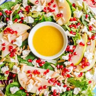A white platter with a mixed greens salad with pears and pomegranates and a citrus dressing in the middle to look like a wreath