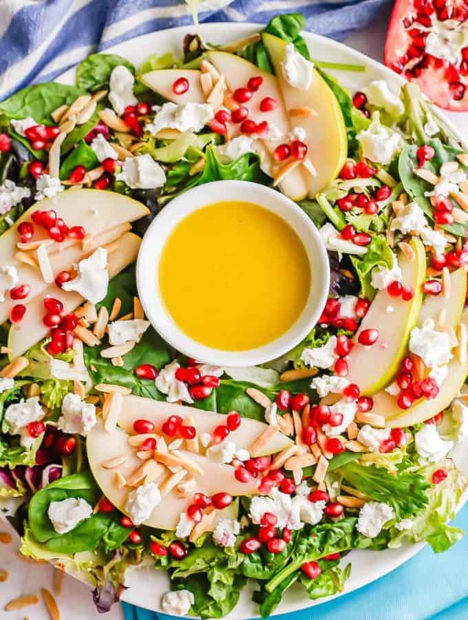 A white platter with a mixed greens salad with pears and pomegranates and a citrus dressing in the middle to look like a wreath