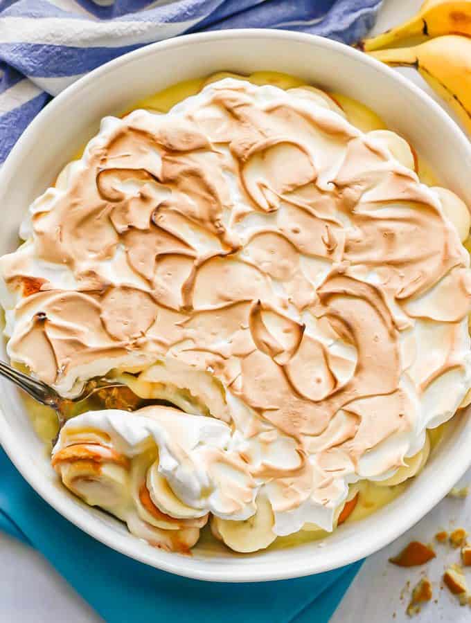 A large white round casserole dish with homemade banana pudding and meringue on top and a scoop being taken out