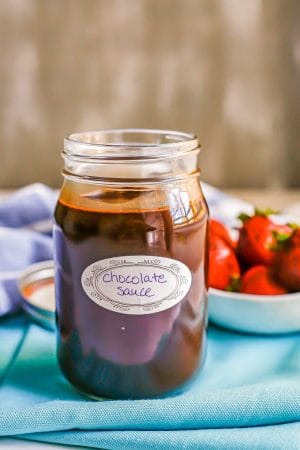 Chocolate sauce in a glass jar with a label and a bowl of strawberries in the background