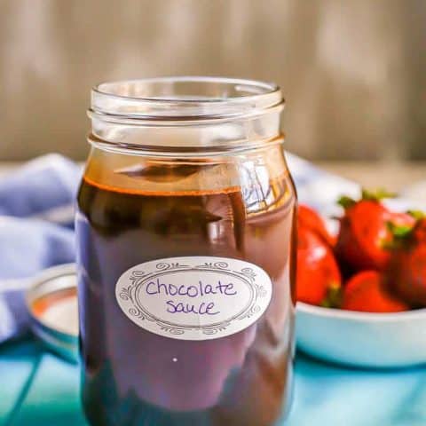 Chocolate sauce in a glass jar with a label and a bowl of strawberries in the background