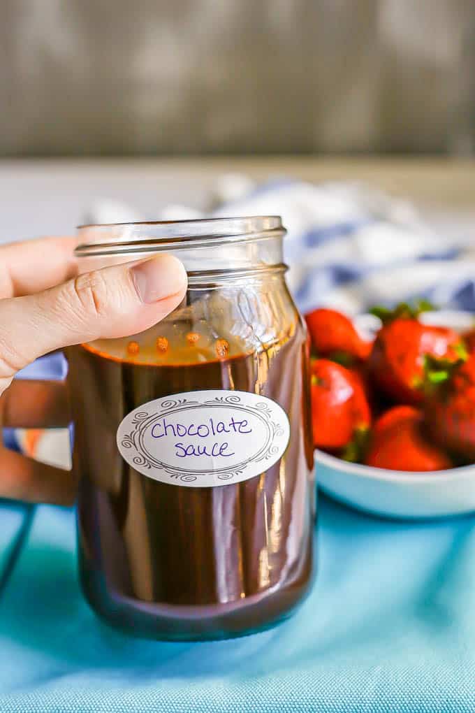 A hand tilting a jar of homemade chocolate sauce and a bowl of strawberries in the background