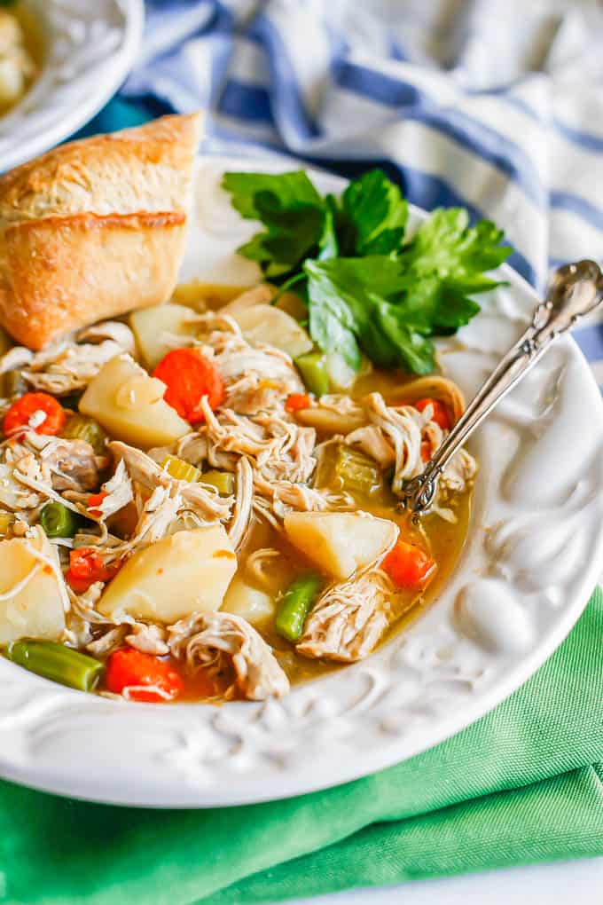 A white soup bowl with chunky chicken stew with parsley and a hunk of bread and a spoon resting in the side